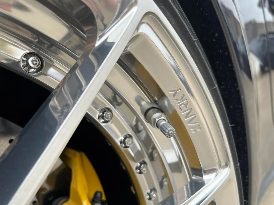 Close-up of a chrome custom wheel with a yellow brake caliper and tire valve, showcasing intricate details.