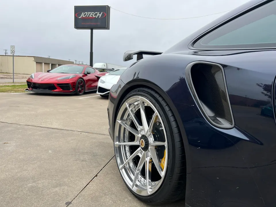 A dark blue Porsche 911 with custom chrome wheels and carbon fiber side vents parked at Jotech Motorsports, with a red Corvette C8 and white Lamborghini Huracan