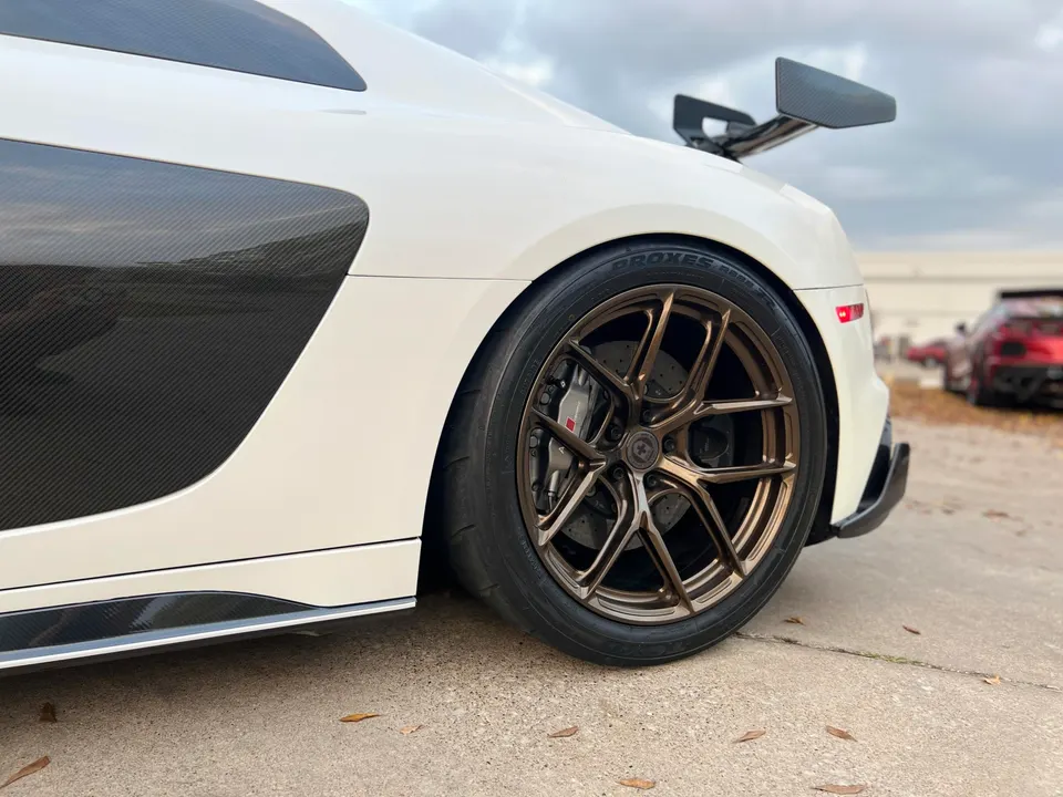 Close-up of a white McLaren's rear wheel with bronze aftermarket wheels, performance brakes, and carbon fiber side panels.