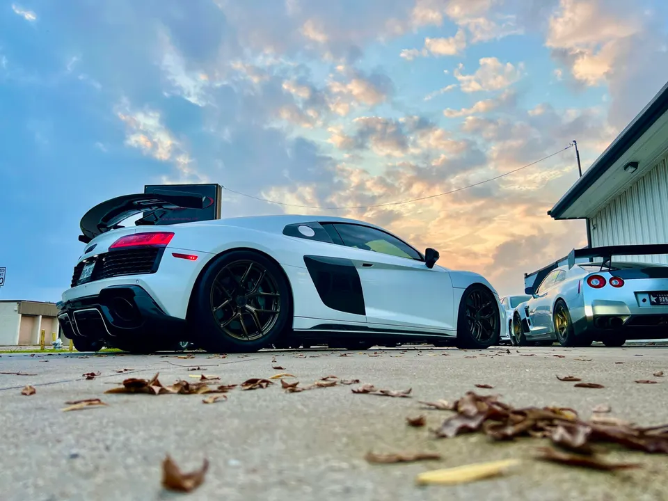 White Audi R8 and silver Nissan GT-R with aftermarket wheels and carbon wings parked outdoors at sunset