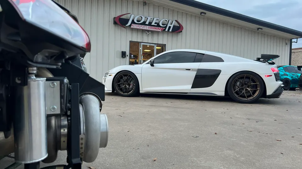 White Audi R8 with custom wheels parked outside Jotech Motorsports, with a large turbocharger visible in the foreground.