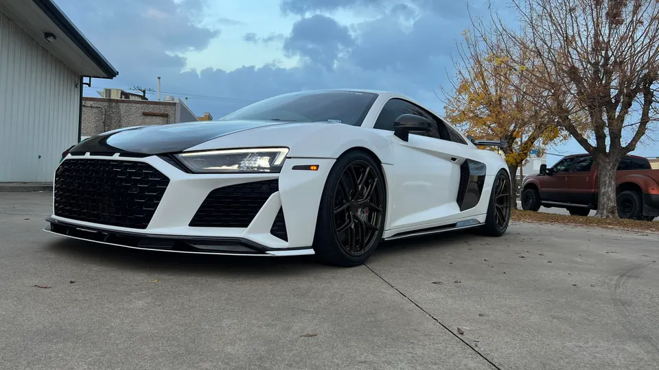 A white Audi R8 with a black hood, custom grille, and front splitter parked outdoors on a cloudy day.