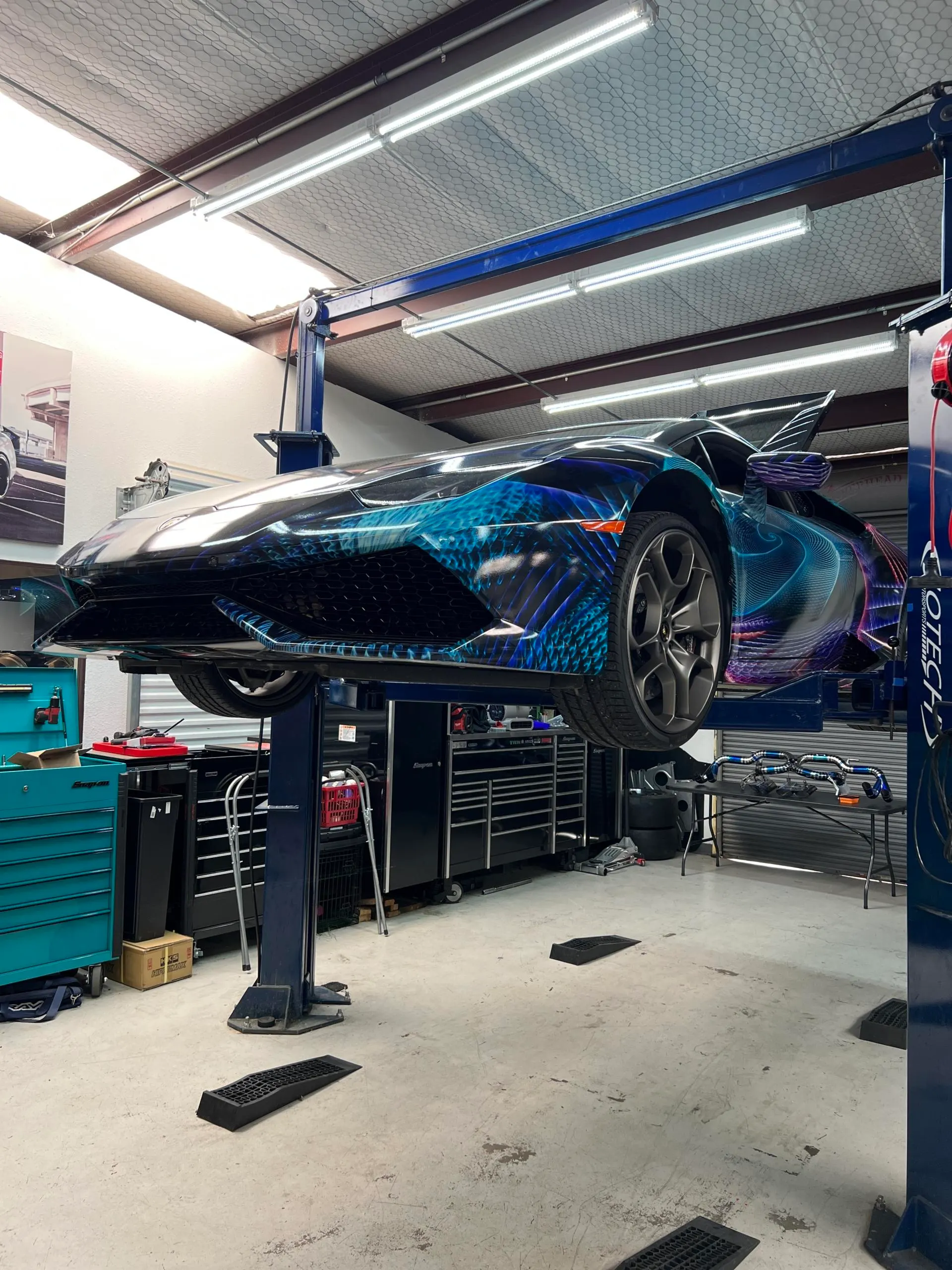 A custom-wrapped Lamborghini Huracan on a car lift inside the Jotech Motorsports performance tuning shop.