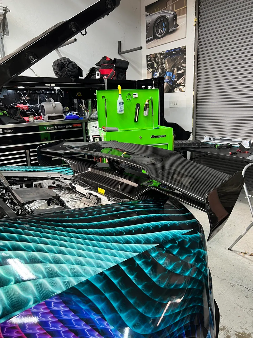 A custom wrapped car with an open trunk and a carbon fiber rear wing being installed in a performance tuning shop.