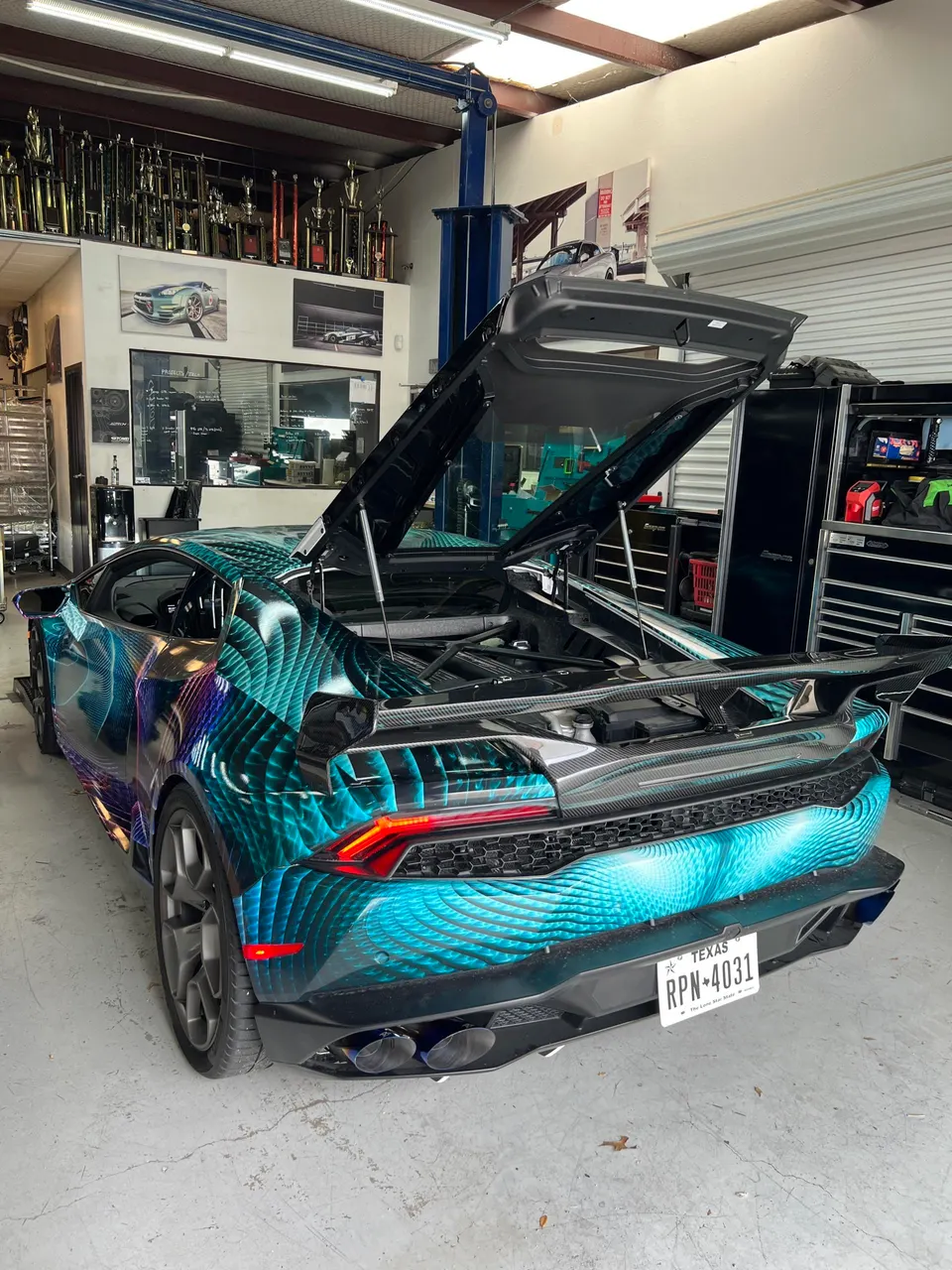A custom-wrapped Lamborghini Huracan with its engine bay open, parked in a performance tuning shop.