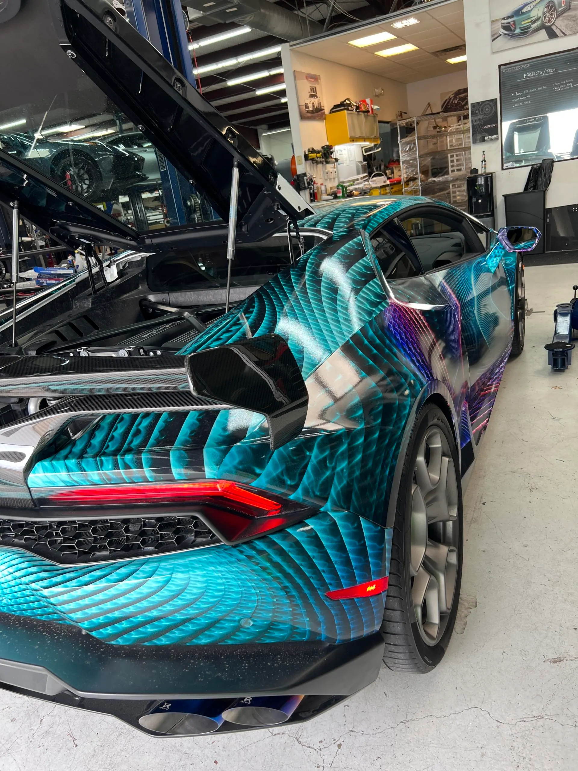 A custom-wrapped Lamborghini Huracan with an open engine bay and aftermarket exhaust in a performance tuning shop.