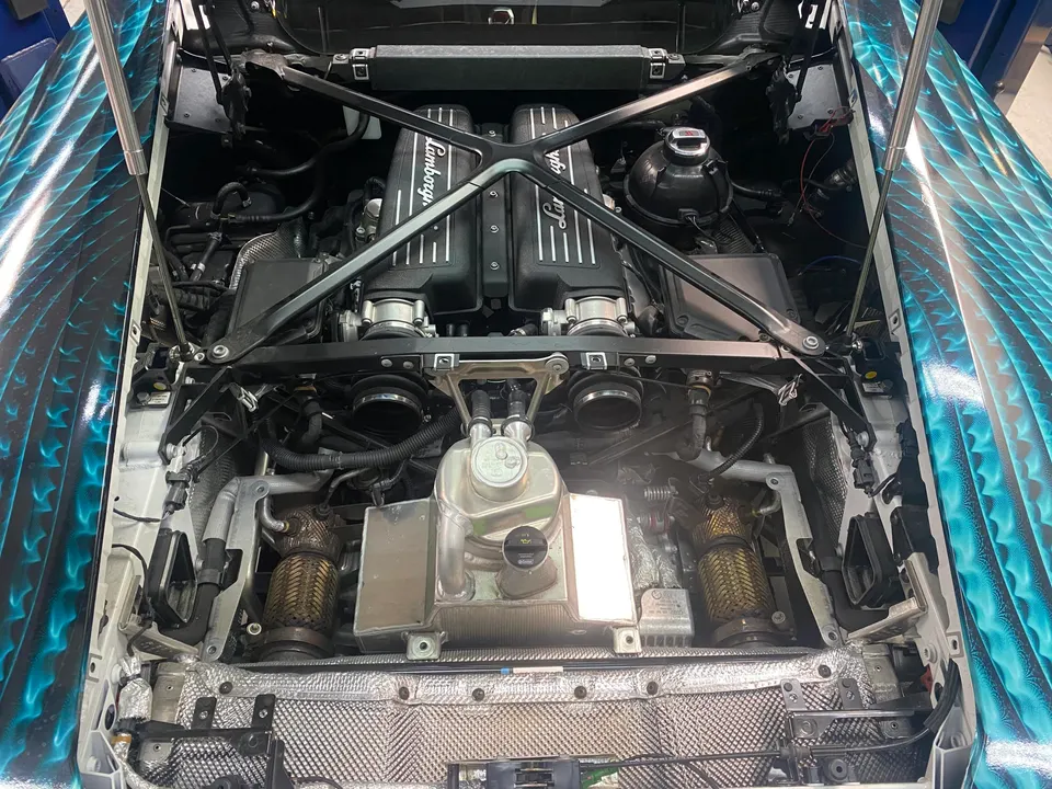 Overhead view of a Lamborghini V10 engine bay with custom blue and black wrap on the hood, showcasing the intake manifolds and exhaust components.