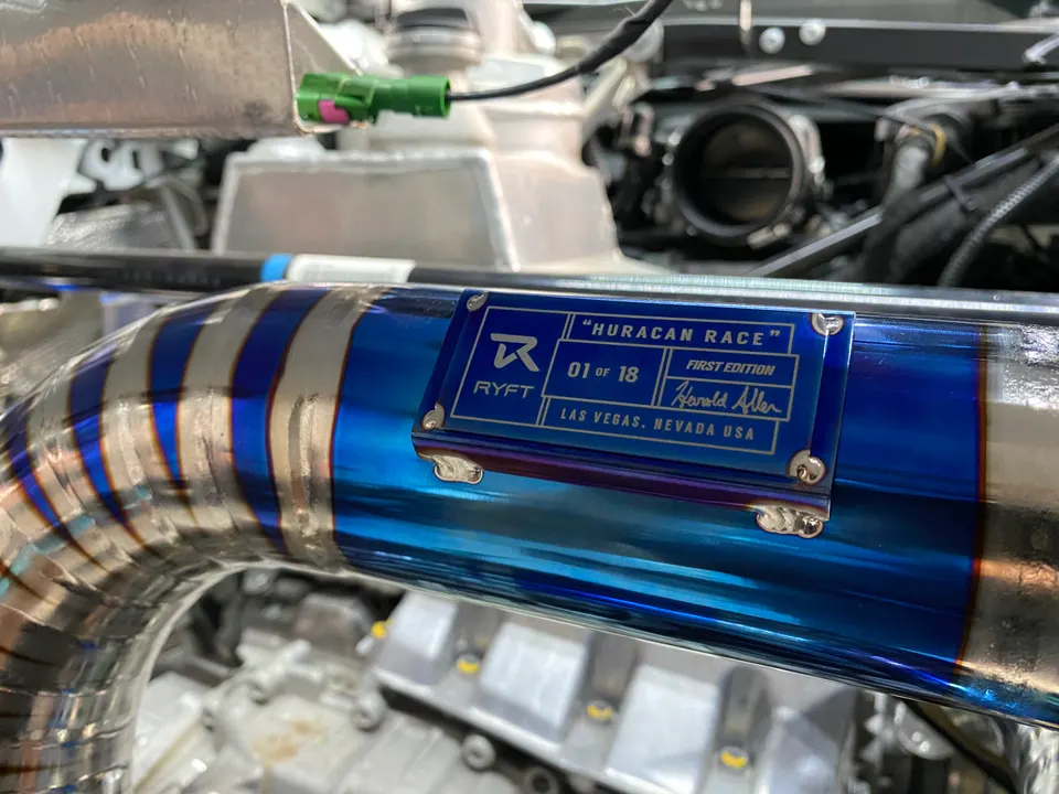 Close-up of a blue-tinted titanium boost pipe with a "RYFT Huracan Race" plaque in a Lamborghini engine bay.