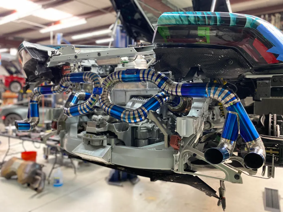 Custom titanium exhaust system being installed on a McLaren supercar in a performance tuning shop, showing intricate piping.