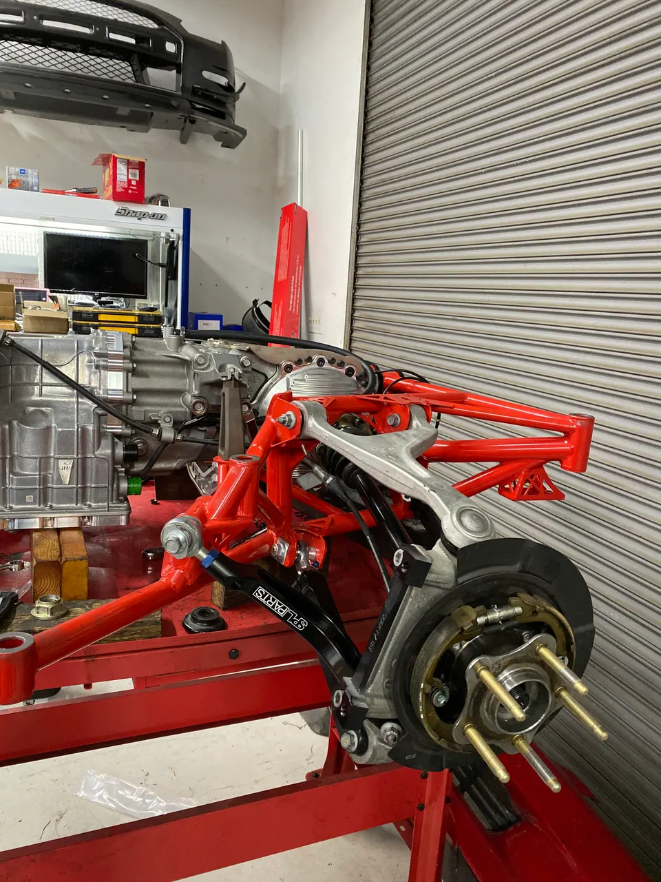 Rear suspension and drivetrain assembly in a performance tuning shop, featuring red control arms and a silver transaxle.