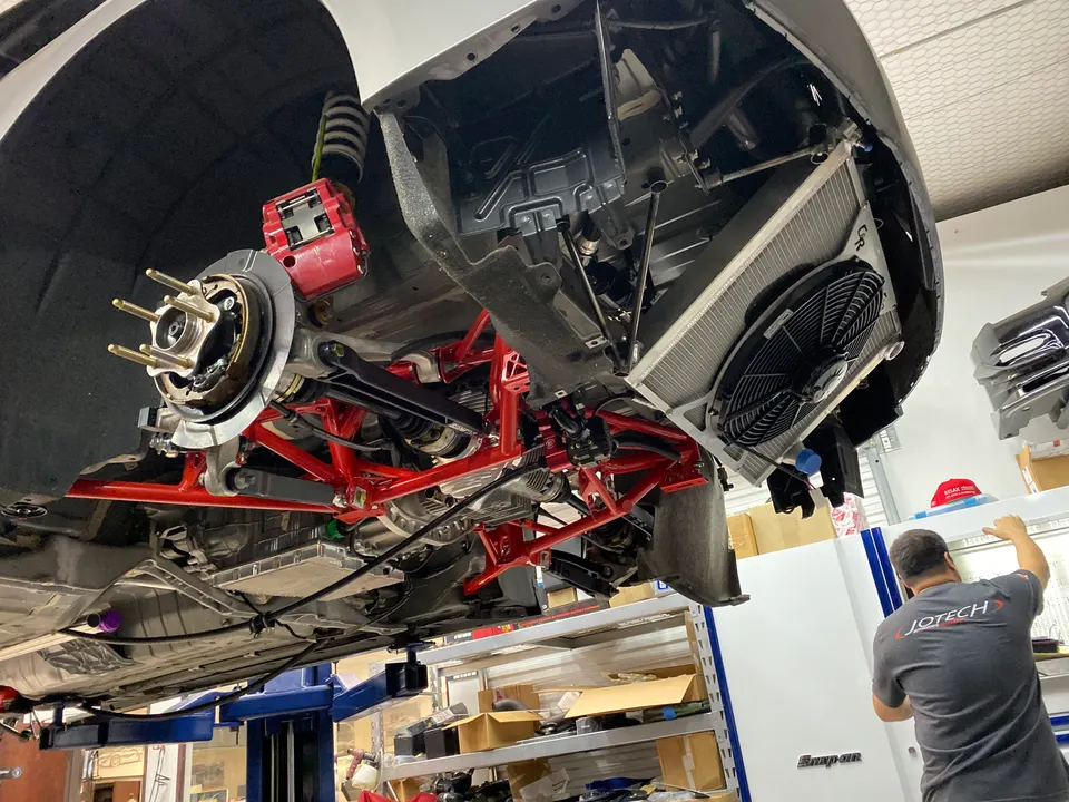 Underside view of a car on a lift in a shop, showcasing red suspension components, brakes, and a large radiator.