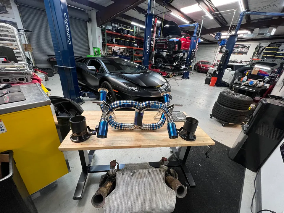 A custom titanium exhaust system on a workbench in a performance tuning shop with a black Lamborghini Aventador in the background.
