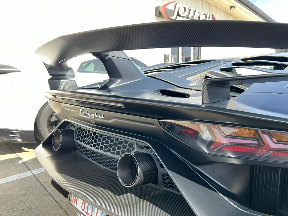 A close-up of the rear of a matte black Lamborghini Aventador with a large carbon fiber wing and dual exhaust tips.