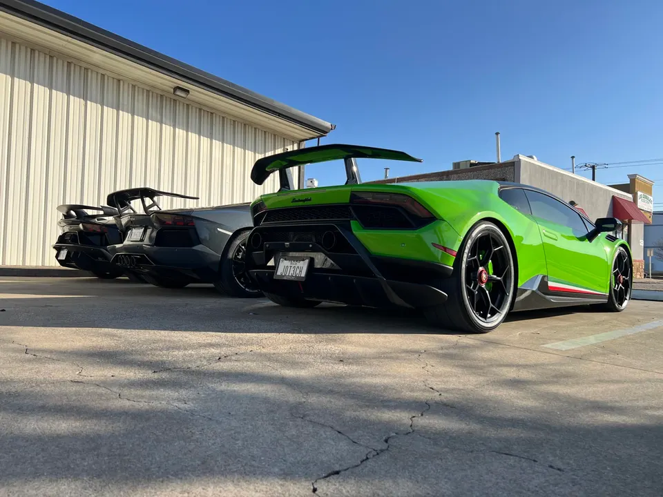 A line of high-performance Lamborghini Huracan and Aventador supercars with large rear wings parked outside.