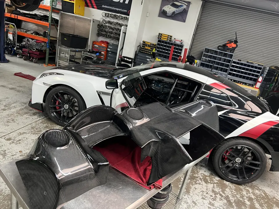 A white and black Nissan GT-R in a performance shop with carbon fiber exterior aero parts laid out on a table.