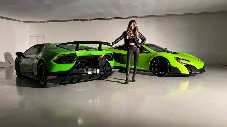 A woman in a black leather jacket stands between a green Lamborghini Huracan and a green McLaren supercar in a studio.