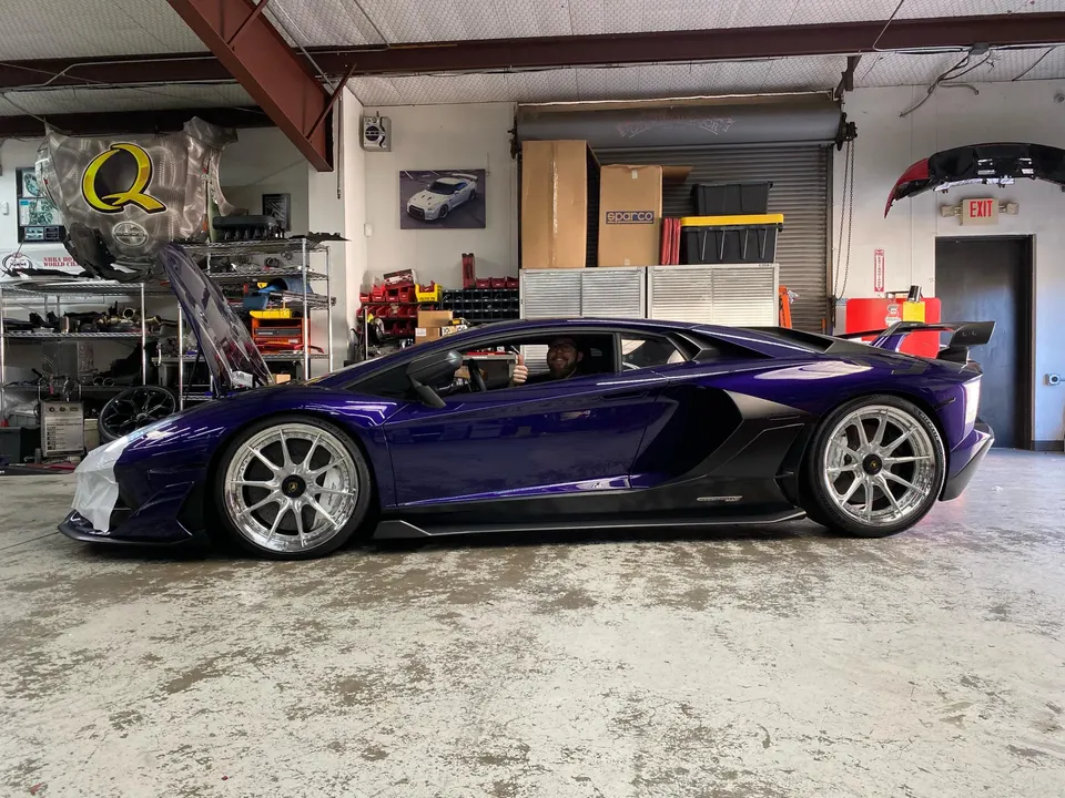 A purple Lamborghini Aventador with custom wheels in a performance tuning shop with its hood open and a person inside.