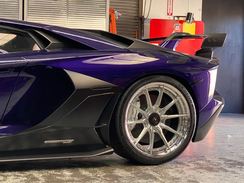 A close-up of a purple Lamborghini Aventador SV's rear wheel, custom silver multi-spoke rim, and carbon fiber wing.