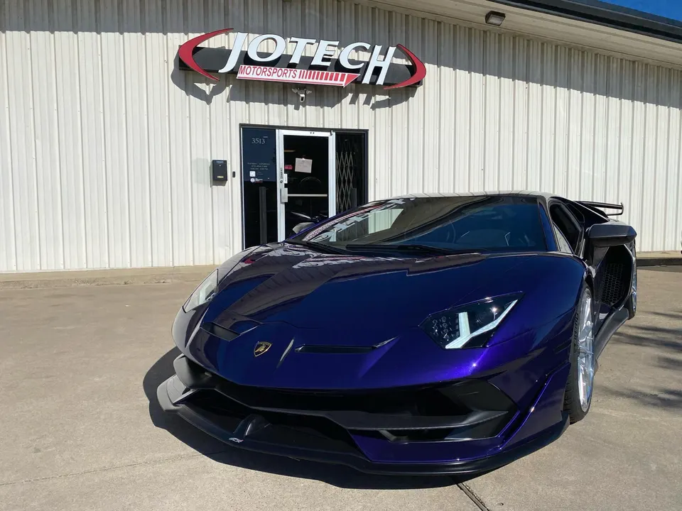 A purple Lamborghini Aventador parked in front of the Jotech Motorsports building on a sunny day.