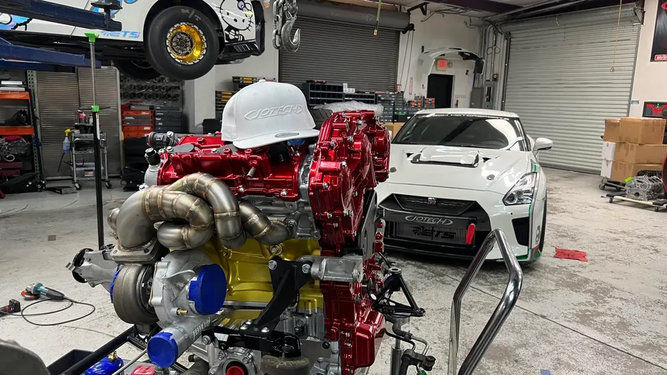 A Jotech Motorsports built GT-R engine with custom turbos and exhaust manifolds in a performance shop, with a white GT-R in the background.