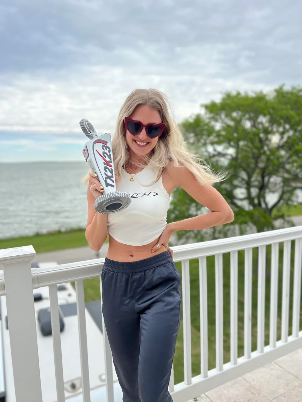 A smiling woman in heart-shaped sunglasses holds a TX2K23 trophy on a balcony overlooking water and trees.