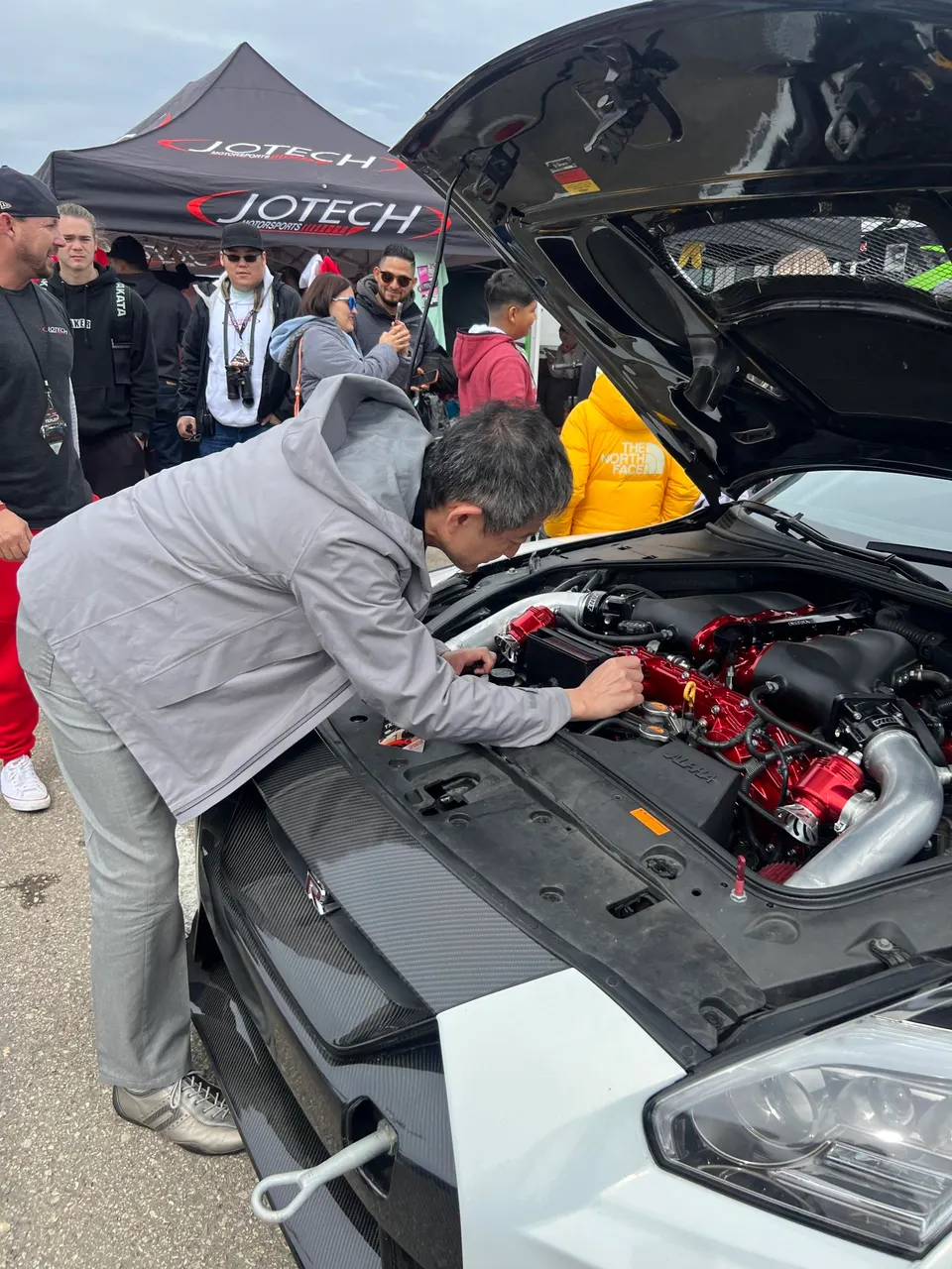 Man inspecting the modified engine bay of a white Nissan GT-R at a Jotech Motorsports event, showcasing aftermarket induction parts.
