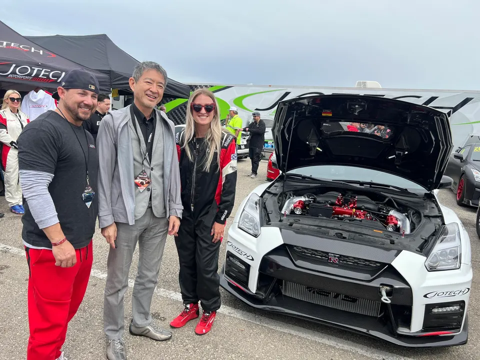 Three people smiling next to a white Nissan GT-R with its hood open, showcasing a modified engine at an outdoor event.