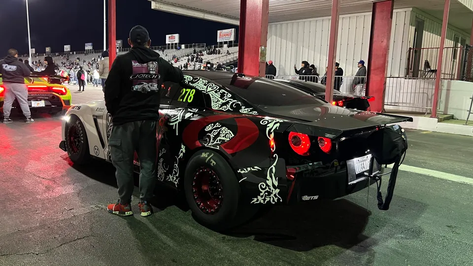 A custom black and white wrapped Nissan GT-R with drag slicks and a parachute at a drag strip at night, with a person standing nearby.