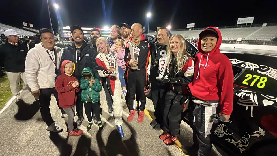 A group of people, including children, celebrating with trophies at a night drag racing event next to a race car.