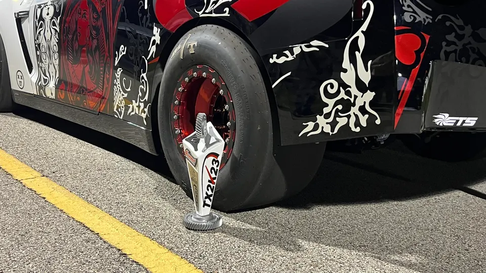 A Nissan GT-R drag car with custom wheels and a drag slick tire, with a TX2K23 trophy on the asphalt.