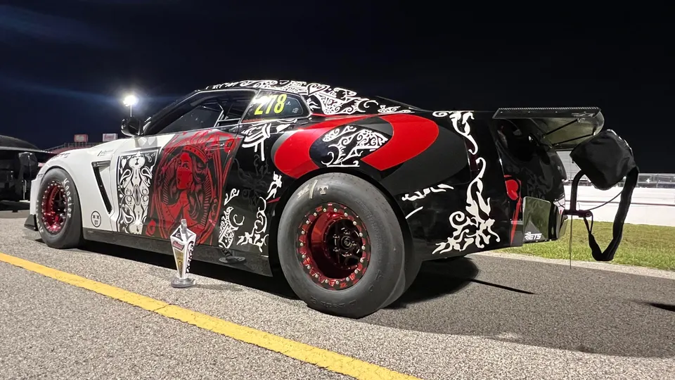 A Nissan GT-R with a custom black, white, and red livery, drag slicks, and a parachute, parked on a drag strip at night with a trophy.