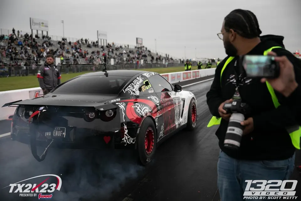 Custom wrapped Nissan GT-R R35 performing a burnout on a drag strip with a large rear wing and red wheels.