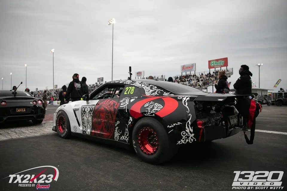 A black, white, and red custom-liveried Nissan GT-R with drag wheels and a parachute at a drag strip during TX2K23, with another GT-R in the background.