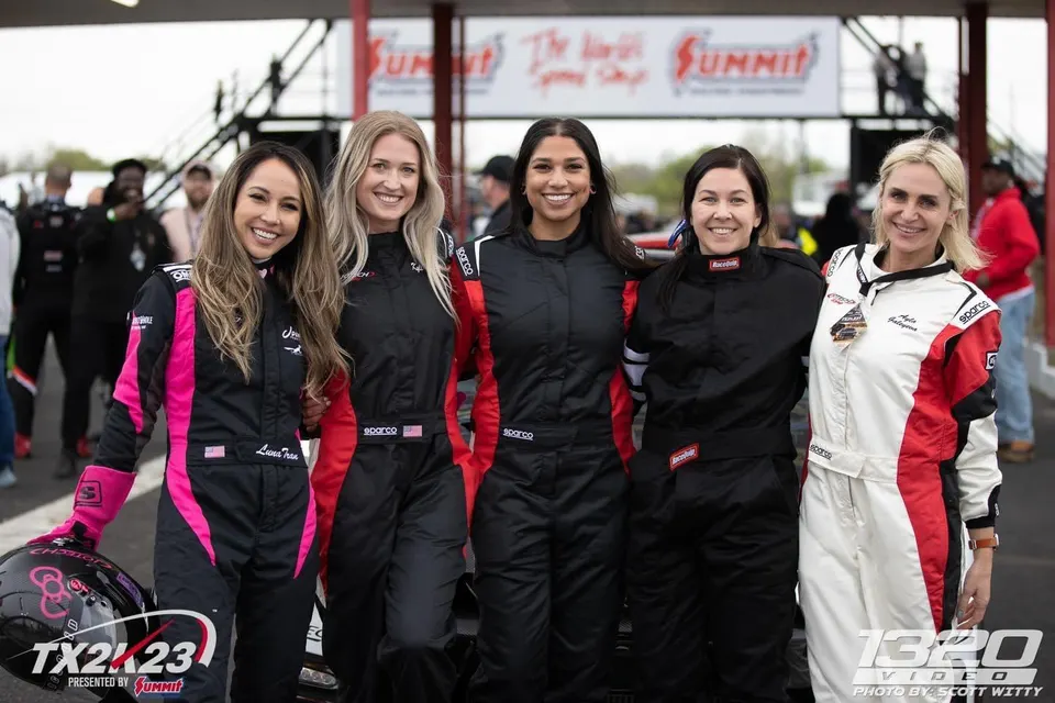 Five women in racing suits pose for a photo at a drag strip event, one holding a racing helmet.