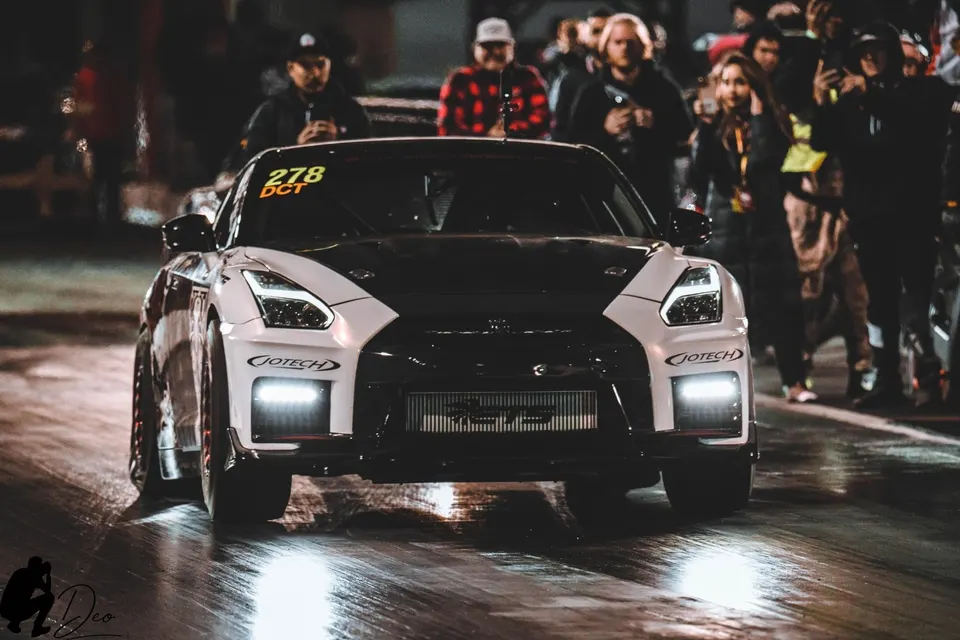 A white and black Nissan GT-R with race number 278 on a drag strip at night, with spectators in the background.