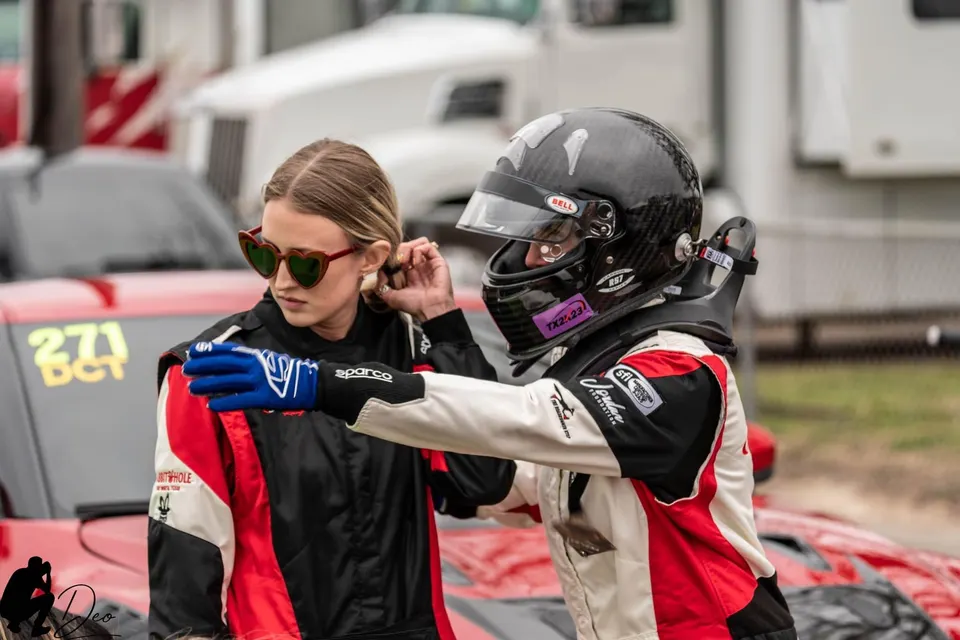 Two racers in full gear, one wearing a helmet and the other heart-shaped sunglasses, standing next to a red drag car at the track.