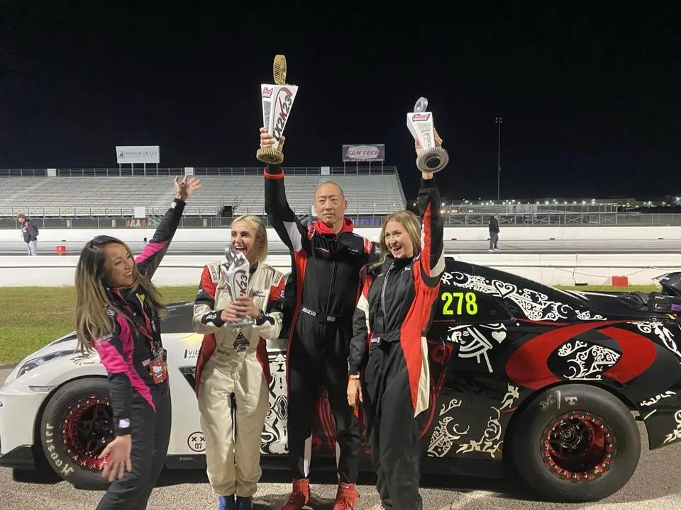 Racers celebrate with trophies at a night drag race event, standing next to a white and a black Nissan GT-R.