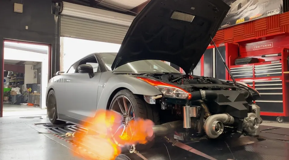 A silver Nissan GT-R on a dyno with its hood open, shooting flames from the exhaust during a high-horsepower run, showcasing large turbochargers.