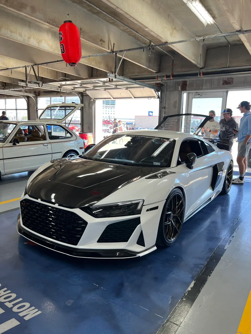 White Audi R8 with a carbon fiber hood and front aero parked in a garage with people around.