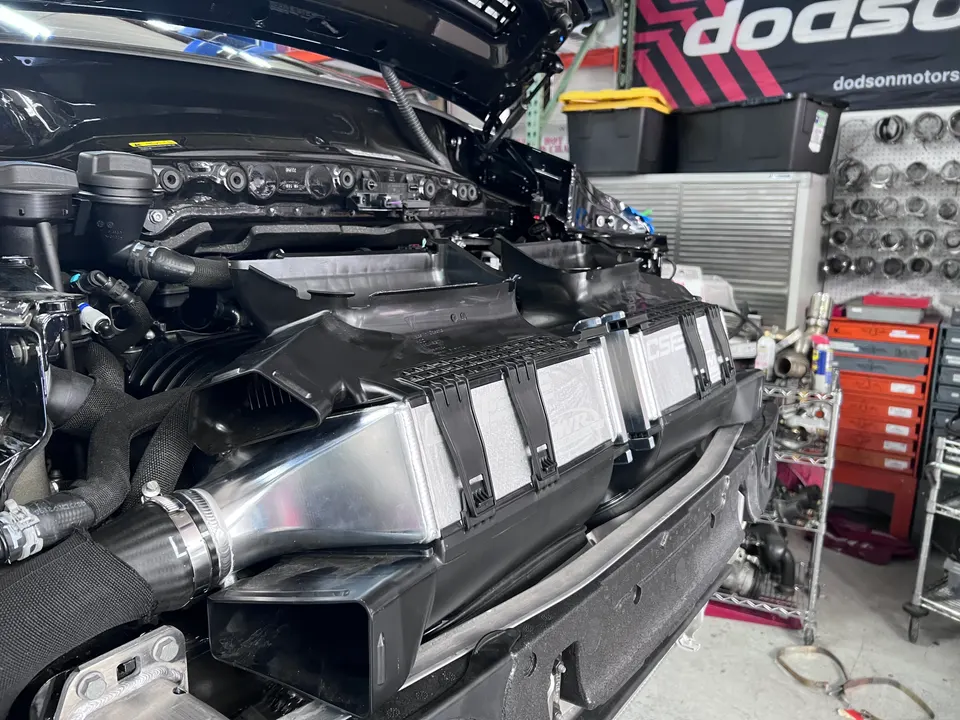 Front view of a car in a shop with the bumper removed, showcasing a large CSF intercooler and boost piping during installation.