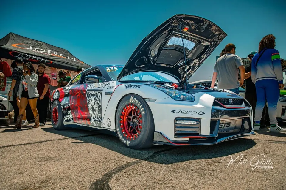 Highly modified white and red Nissan GT-R with hood open at an outdoor car show with a Jotech tent and spectators.