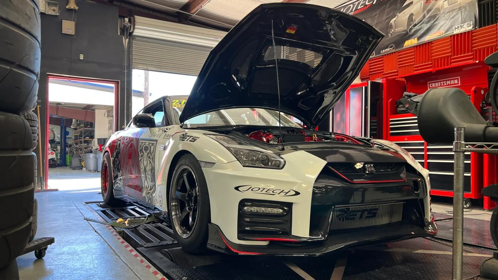 A white Nissan GT-R with its hood open, strapped to a dyno at Jotech Motorsports, showcasing its performance modifications.