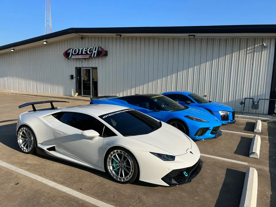 Three high-performance cars, a white Lamborghini Huracan, blue Corvette C8, and blue Audi R8, parked outside Jotech Motorsports shop.