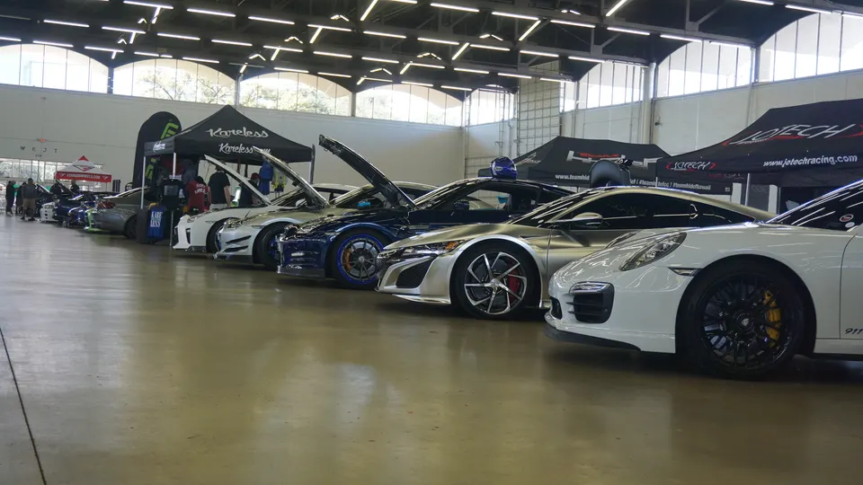 A line of modified sports cars, including an Acura NSX, Porsche 911, and Nissan GT-R, at an indoor car show.