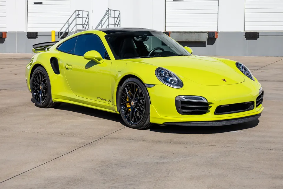 Bright yellow Porsche 911 Turbo S parked outdoors in front of a white industrial building on a sunny day.