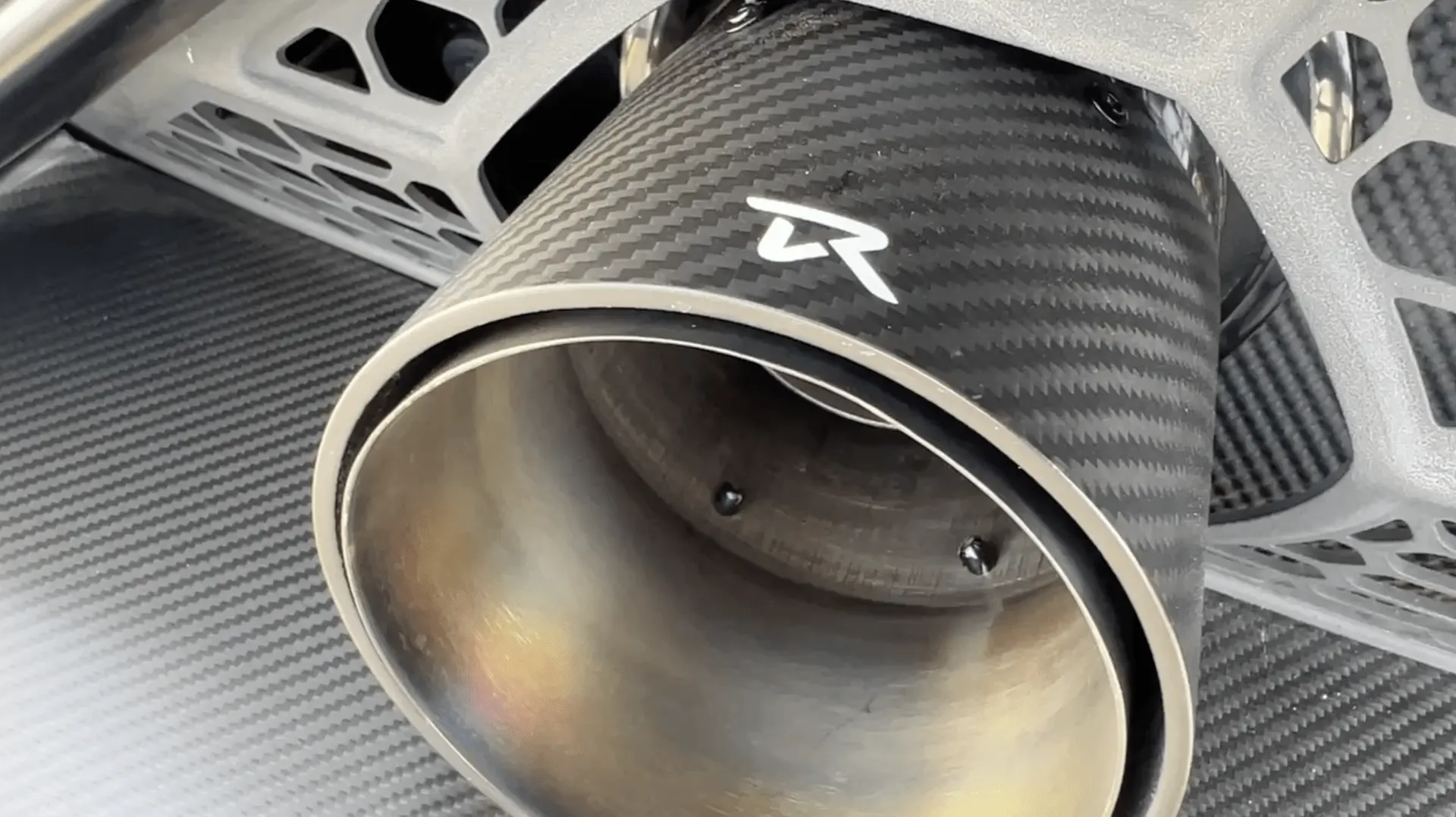 Close-up of a carbon fiber exhaust tip with a logo, showcasing automotive detail and texture.