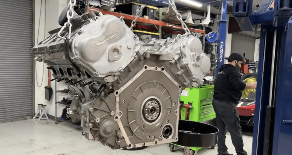 A large V8 engine suspended by chains in a performance tuning shop, with a mechanic working nearby.