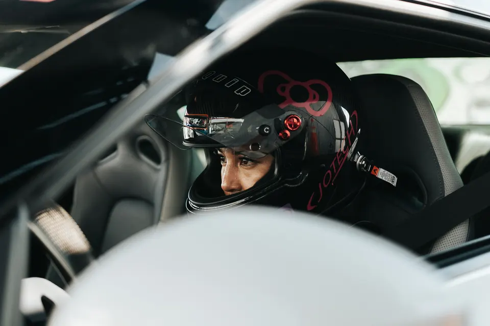 A driver wearing a black racing helmet with a pink Hello Kitty decal sits in the interior of a performance car.