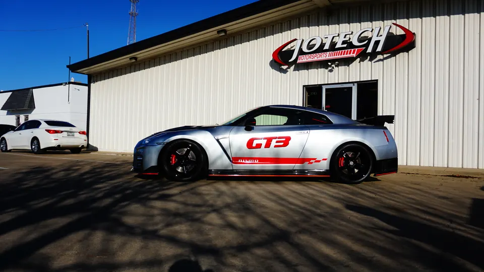 A silver Nissan GT-R R35 with GT3 livery parked outside Jotech Motorsports performance shop on a sunny day.
