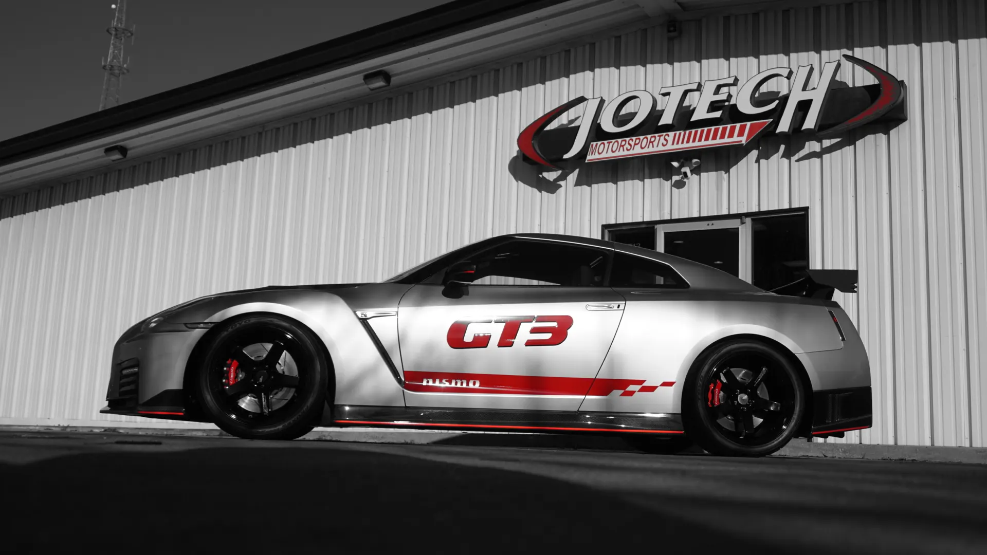 Silver Nissan GT-R with GT3 livery parked outside Jotech Motorsports shop building, showcasing the car's profile.